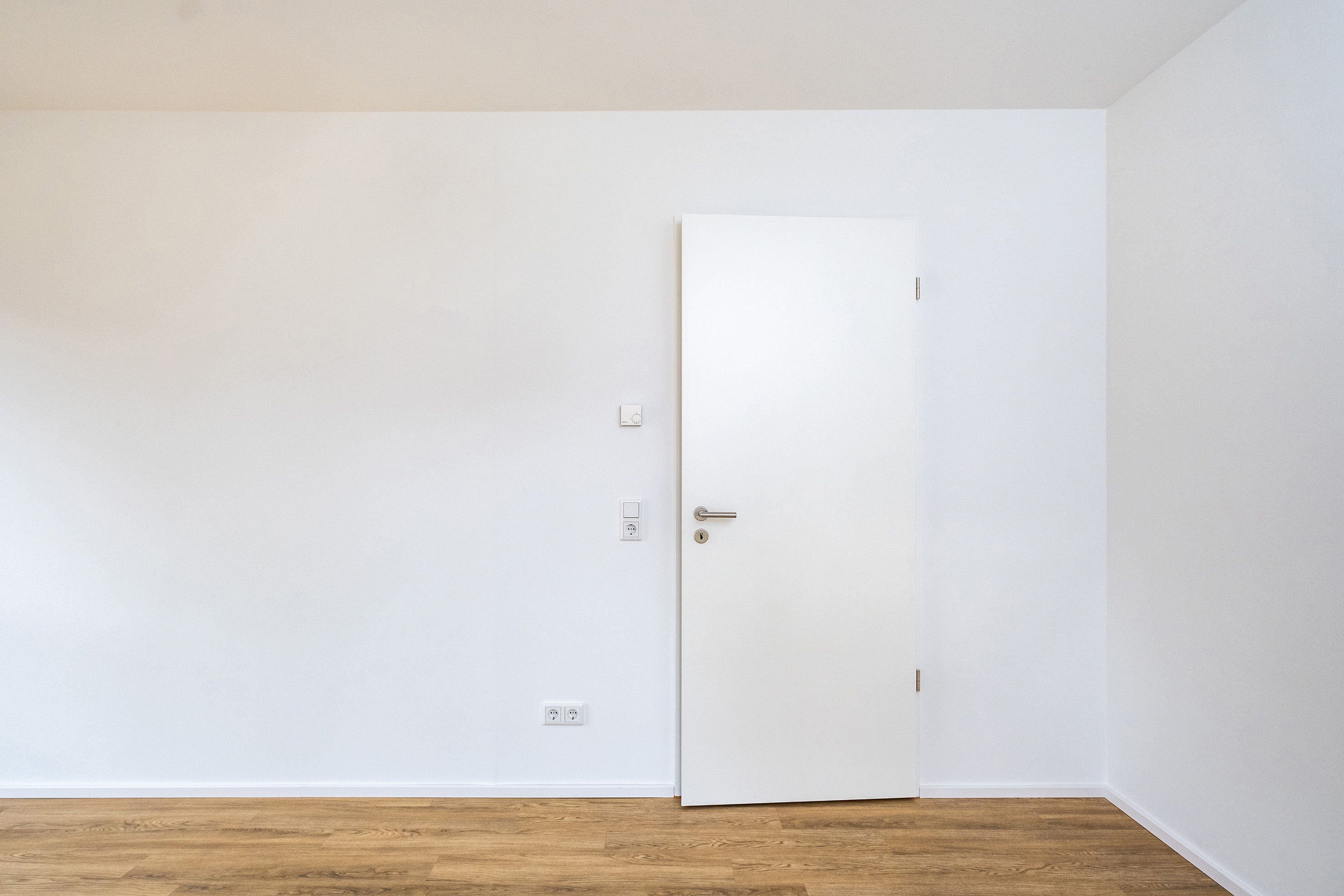 Neubau: ein Zimmer mit angelehnter Tür, alle Wände sind weiß, der Boden ist aus Holz